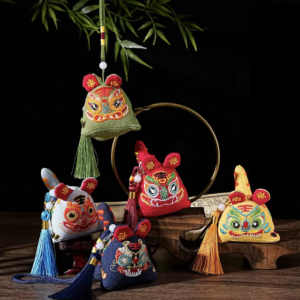 Five detailed fabric sachet pendants, handcrafted in the shape of traditional Chinese tiger heads, are displayed together on a dark wooden stand. Each tiger is intricately embroidered with colorful threads of red, orange, green, blue, and gold. Their faces feature wide-set eyes, stylized noses, whiskers, and small "Wang" characters for "King" on their foreheads. Each tiger is a different color: soft green, sky blue, navy blue, ruby red, and golden yellow. All but the navy one hang from coordinating cords and end in large, silky tassels matching the sachet's color, weighted with round white beads. The top sachet, green, hangs from a green cord attached to a cluster of leafy green bamboo. In the background, there is a round bronze mirror frame and open Chinese texts with brush script, with everything illuminated by warm light against a dark background.